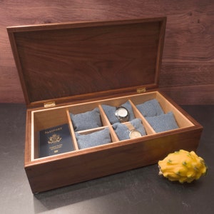 May include: A wooden watch box with a hinged lid, open to reveal six compartments lined with grey fabric. Two watches are visible in the compartments, along with a passport. The box is made of dark wood with a natural grain pattern.