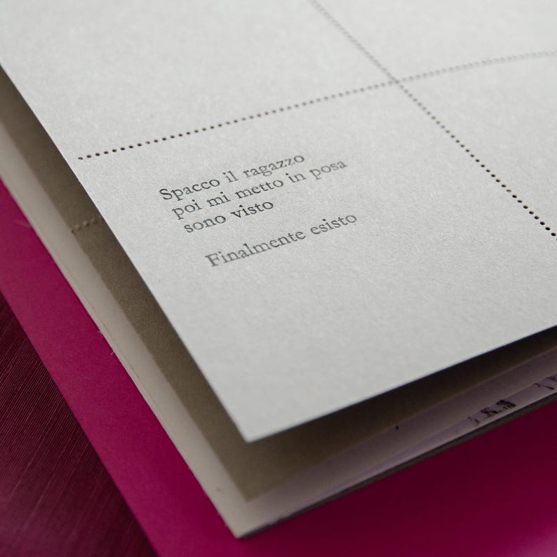 May include: A close-up of a gray notebook with a dotted line border. The notebook has the following text printed on it: "Spacco il ragazzo poi mi metto in posa sono visto finalmente esisto"