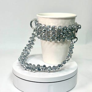 May include: A white paper cup with a silver beaded handle and band. The band and handle are made of small, faceted beads strung together. The cup is sitting on a white, circular platform.