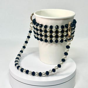 May include: A white paper cup with a beaded cup holder and strap. The cup holder is black and white, with gold-coloured clasps. The strap is made of alternating black and white beads. The cup is on a white platform.