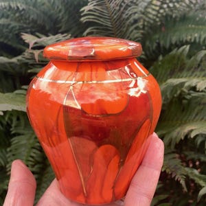 May include: Orange ceramic cremation urn with a gold trim and a lid. The urn has a marbled design with streaks of gold.