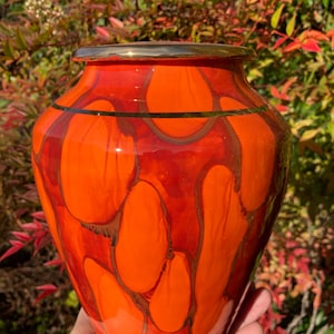 May include: A vibrant orange and red ceramic vase with a glossy finish. The vase features a gold rim and a pattern of organic shapes in shades of orange and red. The vase is held in front of a background of green and red foliage.