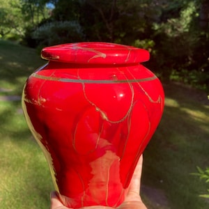 May include: A bright red ceramic urn with a matching lid, featuring a glossy finish and irregular, thin gold-colored veins that run vertically across the surface. The vessel has a rounded body that tapers toward the base and a wide opening.
