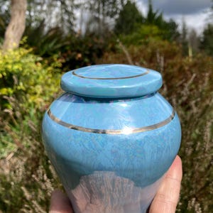 May include: A small, light blue ceramic urn with a gold trim around the top and bottom. The urn has a marbled effect and a glossy finish.