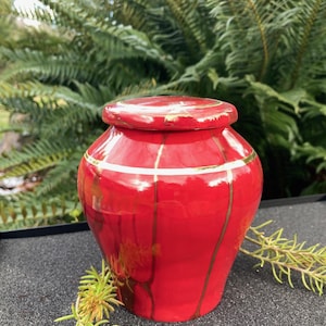 May include: A red ceramic urn with gold accents. The urn has a smooth, glossy finish and a rounded shape.