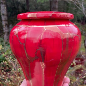 May include: A red ceramic urn with gold accents and a marbled design. The urn has a lid and is perfect for storing ashes.