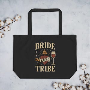 May include: Black tote bag featuring the text "BRIDE LOVE TRIBE" in a vintage font. The design includes a flaming heart with a banner reading "LOVE" and a wine glass. The bag has two black handles.