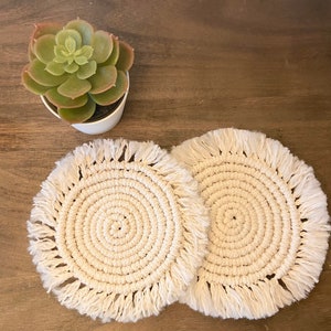 May include: Two white crocheted coasters with fringe edges. The coasters are round and have a circular pattern in the center.