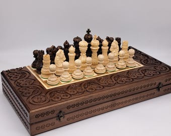 May include: A wooden chess set with intricately carved details. The dark brown wooden box holds a chessboard with ivory and dark brown chess pieces. The set is closed with a metal clasp.
