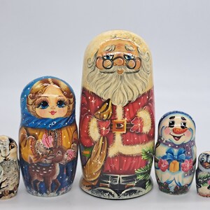 Peut inclure: Un ensemble de cinq poupées russes traditionnelles, ou poupées gigognes, représentant un Père Noël, une femme avec un renne, un bonhomme de neige et deux poupées plus petites. Les poupées sont peintes dans des couleurs vives et présentent des détails complexes.