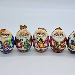 Puede incluir: Cinco adornos de madera pintados a mano con forma de huevo, cada uno representando a un Papá Noel diferente con barba blanca y gafas. Los adornos están decorados con varios temas navideños, incluyendo un pájaro, una casa y una campana. Cada adorno tiene una cinta dorada para colgar.
