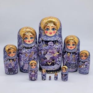 May include: A collection of Matryoshka dolls, or nesting dolls, in shades of lavender and gold. The dolls depict winter scenes and have painted faces with blue eyes and golden headdresses. The set includes dolls of different sizes, from large to small.