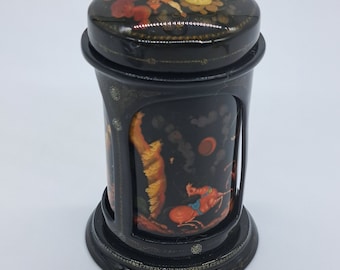 May include: A black lacquered cylindrical box with a hinged lid. The box is decorated with intricate gold and silver details and features a colorful scene painted on the lid and sides. The scene depicts a group of figures in a whimsical setting.