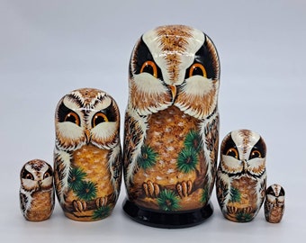 May include: Set of five nesting Russian dolls, each painted with a brown and white owl with orange eyes. The owls are surrounded by green pine needles and branches. The largest doll is about 15 cm tall.