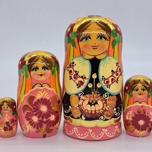 May include: A set of five traditional Russian nesting dolls, or matryoshka dolls. The dolls are painted in a colourful floral pattern with pink, orange, yellow, green, and black accents. The largest doll is wearing a black and white patterned dress with a floral design. The dolls are all smiling and have brown hair.