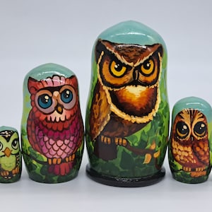 Pode incluir: Conjunto de cinco bonecas russas aninhadas, cada uma pintada com um design de coruja diferente. A boneca maior é marrom com olhos amarelos, a próxima é rosa com olhos azuis, a próxima é verde com olhos amarelos, e as duas menores são marrons com olhos amarelos.