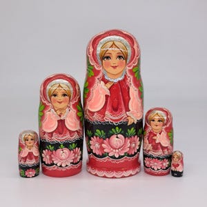 May include: A set of six Russian nesting dolls, or Matryoshka dolls, in varying sizes. Each doll features a painted face and is dressed in a red and pink outfit with floral and silver accents. The dolls are arranged in a row.