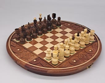 May include: A circular wooden chess set featuring a square board with alternating light and dark brown squares. The board is surrounded by a wide, carved wooden border with geometric and floral patterns. Dark and light wooden chess pieces are arranged.