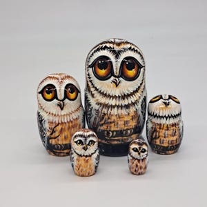 Könnte beinhalten: Ein Satz von fünf Holz-Matrjoschka-Puppen, die jeweils einer Eule nachempfunden sind. Die Puppen sind in Braun-, Weiß- und Orangetönen bemalt. Die größte Puppe befindet sich in der Mitte, die kleinste unten. Die Puppen sind unterschiedlich groß.