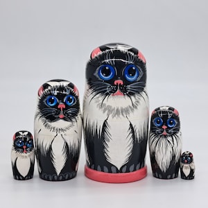 Pode incluir: Um conjunto de cinco bonecas russas de encaixe pretas e brancas, cada uma representando um gato com olhos azuis. A maior boneca tem cerca de 25 cm de altura e a menor cerca de 5 cm.