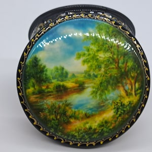 以下が含まれることがあります： 黒地に金色の縁取りが施された丸い木製の箱。蓋には、青い空の下、緑の森林を流れる川が描かれた絵が描かれています。