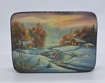 May include: A hand-painted wooden box with a winter scene. The box depicts a snowy landscape with a small village, a frozen river, and a few birds flying overhead. The sky is a soft blue with hints of pink and orange.