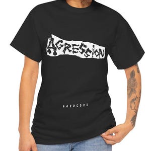 May include: Black t-shirt with the word "AGGRESSION" in a distressed white font across the chest. Below the word "AGGRESSION" is the word "HARDCORE" in white font. The shirt is short-sleeved.