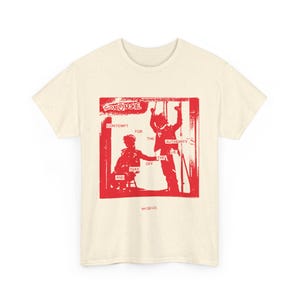 May include: Cream-colored t-shirt with a red graphic print. The graphic features two figures and text that reads "CONDUSE", "CONTEMPT FOR THE AUTHORITY", "TAKE OFF THE LIE", and "AND".