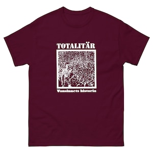 Könnte beinhalten: Ein weinrotes T-Shirt mit einem weißen Aufdruck. Der Aufdruck zeigt das Wort "TOTALITÄR" über einem Schwarz-Weiß-Bild einer Menschenmenge, mit dem Text "Vansinnets historia" darunter. Das Shirt ist aus weichem Material.