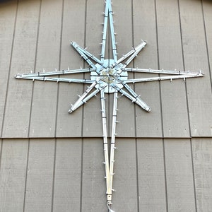May include: A large, outdoor Christmas star decoration with white lights. The star is mounted on a light brown wooden wall. The star has a central square and radiating points, each with small, white lights. A white power cord hangs below.