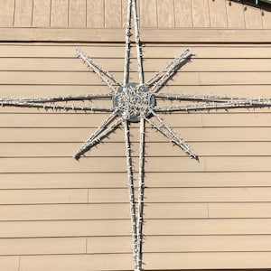May include: A large white star-shaped string of lights is attached to the exterior of a light brown wooden house. The star is made of white lights and has a circular center.