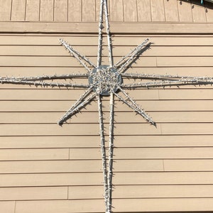 May include: A large, white star-shaped Christmas light decoration mounted on a tan-colored building. The star is made of multiple strands of white lights, creating a bright, festive display. The building has horizontal siding and a row of colorful lights along the roofline.