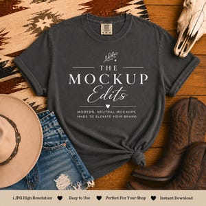 May include: A dark gray t-shirt with the text "THE MOCKUP Edits" in white, along with the tagline "MODERN, NEUTRAL MOCKUPS MADE TO ELEVATE YOUR BRAND." The shirt is styled with denim shorts, a cowboy hat, and boots, suggesting a Western theme.