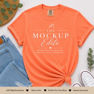 May include: An orange t-shirt with rolled-up sleeves and a knotted hem. The shirt features the text "THE MOCKUP Edits" in white script. The shirt is displayed with blue jeans and white sneakers.