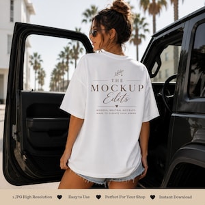 May include: A white t-shirt with the text "THE MOCKUP Edits" in brown, worn by a person. The shirt has a relaxed fit. The background includes a black vehicle and palm trees, suggesting a casual, outdoor setting. The text on the shirt says "MODERN, NEUTRAL MOCKUPS MADE TO ELEVATE YOUR BRAND."