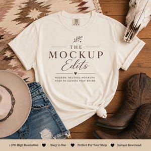 May include: A cream-colored t-shirt with the text "THE MOCKUP Edits" is knotted at the bottom. The shirt is styled with denim shorts, a cowboy hat, and brown cowboy boots. The background includes a patterned rug and a cow skull.