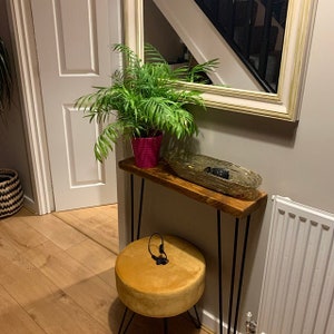 Op de afbeelding: Een houten consoletafel met zwarte haarspeldpoten, een gouden dienblad en een groene plant in een roze pot. De tafel staat in een gang met een gele fluwelen kruk en een witte radiator.