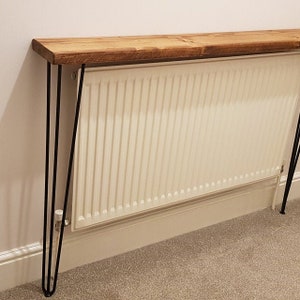 Op de afbeelding: Een houten plank met zwarte haarspeldpoten staat op een witte radiator. De plank is gemaakt van gerecycled hout en heeft een natuurlijke afwerking.