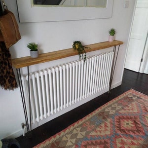 Op de afbeelding: Een houten consoletafel met haarspeldpoten, geplaatst boven een witte radiator. De tafel is versierd met potplanten en heeft een natuurlijke houten afwerking.