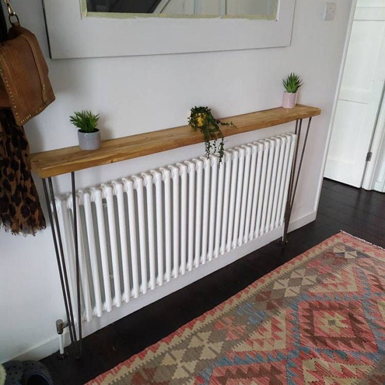 Op de afbeelding: Een houten consoletafel met haarspeldpoten, geplaatst boven een witte radiator. De tafel is versierd met potplanten en heeft een natuurlijke houten afwerking.