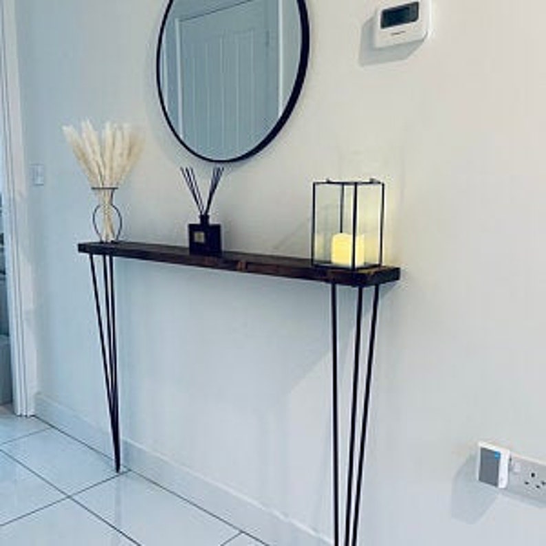 Op de afbeelding: Een donkerbruine houten consoletafel met haarspeldpoten. De tafel is versierd met een vaas met gedroogd pampasgras, een diffuser en een glazen lantaarn.