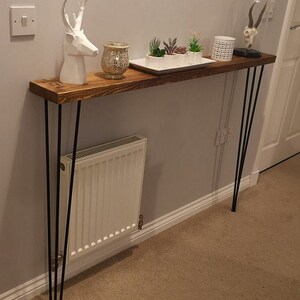 Op de afbeelding: Een rustieke houten consoletafel met een donkerbruine afwerking. De tafel heeft drie zwarte haarspeldpoten en is versierd met een wit hertenbeeldje, een glazen kaarsenhouder en een wit dienblad met kleine potplanten.