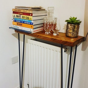 Op de afbeelding: Een houten plank met zwarte haarspeldpoten staat op een witte radiator. De plank is versierd met een stapel kookboeken, een karaf met twee glazen en een potplant met een succulent.