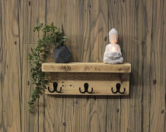 May include: A rustic wooden shelf with three black metal hooks, a small potted plant, and a white Buddha statue holding a candle. The shelf is mounted on a wood panel wall.