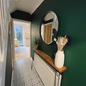 Op de afbeelding: Een gang met een donkergroene muur, een ronde spiegel, een houten consoletafel met zwarte metalen poten en een witte vaas met gedroogde bloemen. De vloer is betegeld met zwart-witte gepatenteerde tegels.