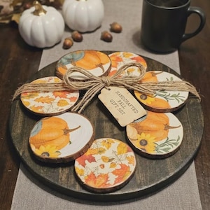 May include: A set of six round wooden coasters featuring autumn-themed illustrations of pumpkins, sunflowers, and fall leaves. The coasters are tied together with twine and include a tag that reads HANDCRAFTED FALL GIFT SET, all arranged on a dark wood tray.