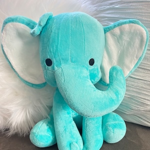 May include: A plush turquoise elephant toy with large ears and a trunk. The elephant has black button eyes and is sitting in front of a white fluffy pillow and a textured silver pillow.