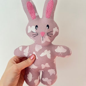 May include: A soft, pink bunny plush toy with embroidered pink ears and white butterfly patterns. The bunny has black eyes and a pink nose.