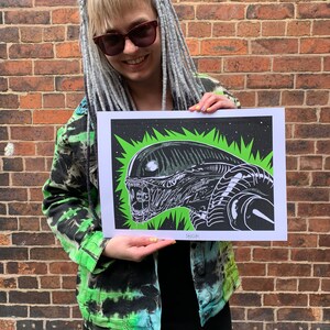 May include: A black and white illustration of an alien creature with a green background. The illustration is printed on a white piece of paper and is being held by a person. The text "SASHISH" is printed in the bottom right corner of the illustration.