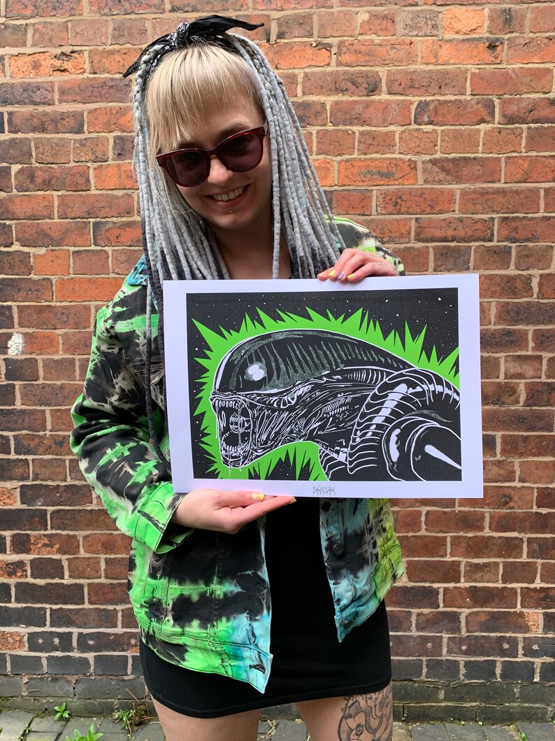 May include: A black and white illustration of an alien creature with a green background. The illustration is printed on a white piece of paper and is being held by a person. The text "SASHISH" is printed in the bottom right corner of the illustration.